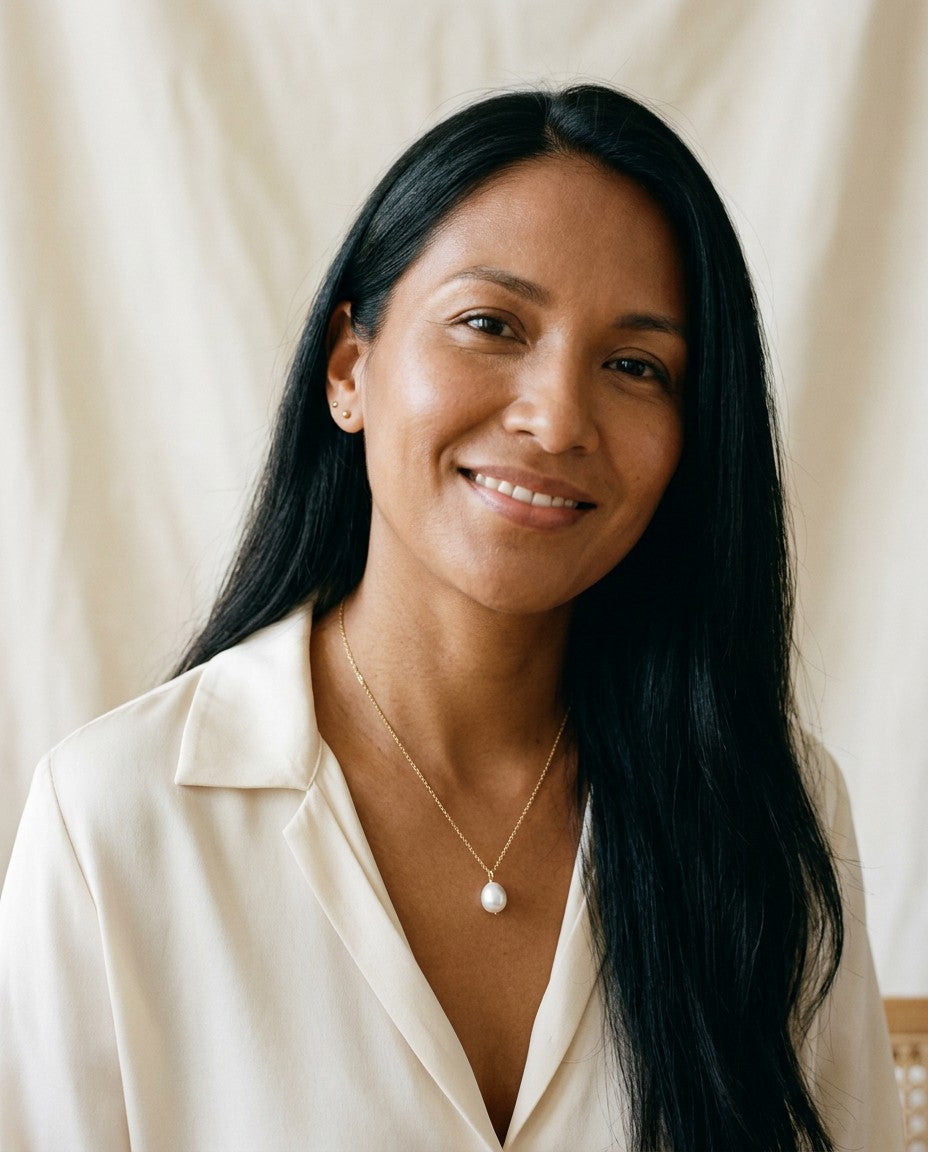 Smiling woman with long black hair in a cream silk blouse wearing a gold necklace with a white pearl drop