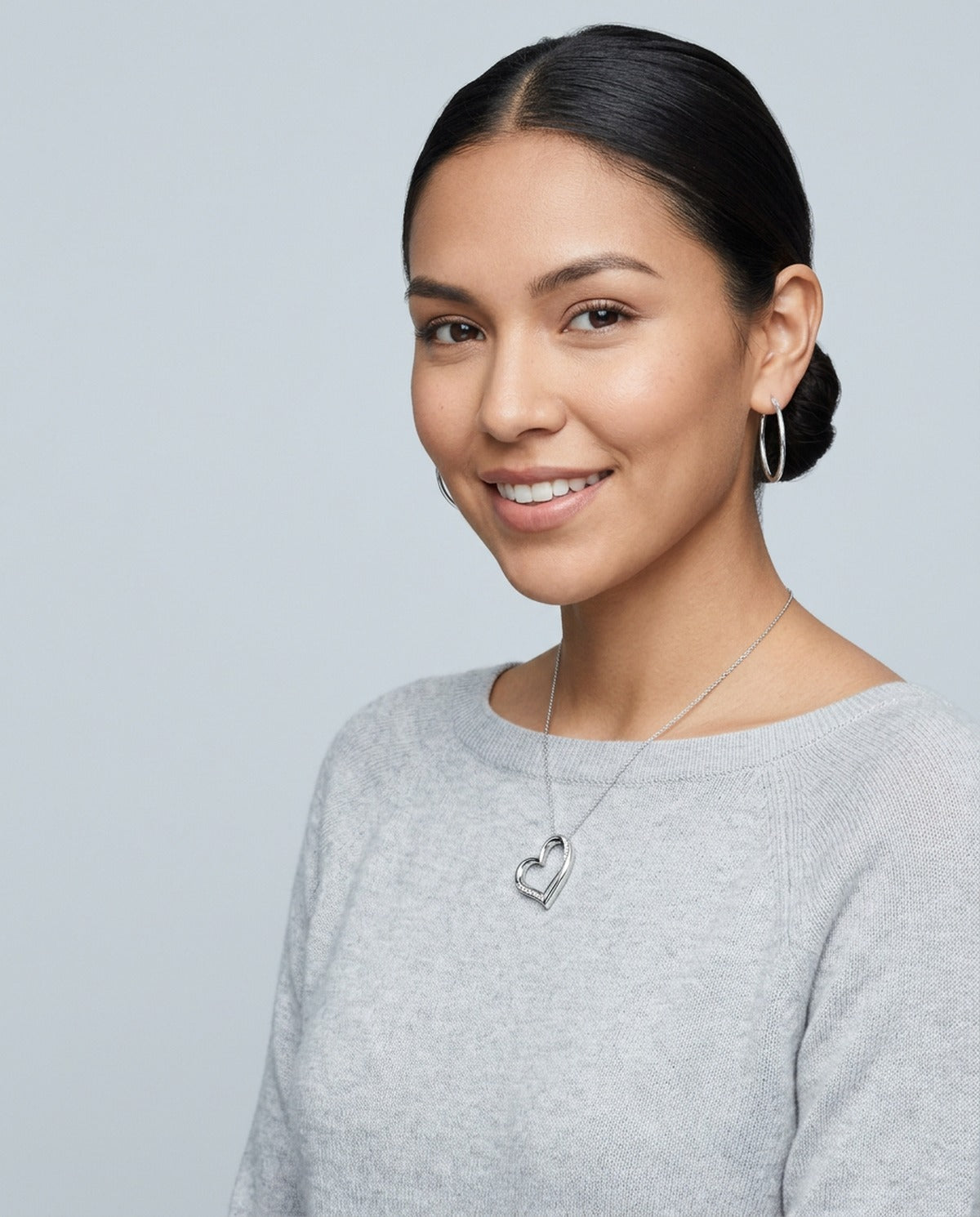 Woman in a gray sweater wearing a sterling silver heart necklace and silver hoop earrings
