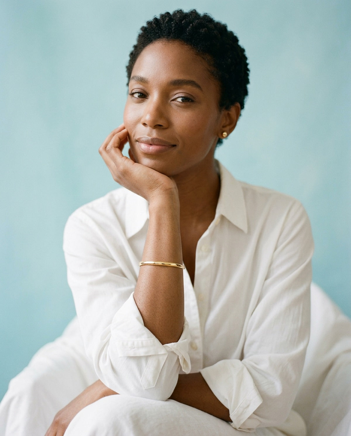 Woman with short natural hair in a white shirt resting her chin on her hand, wearing a slim gold bangle