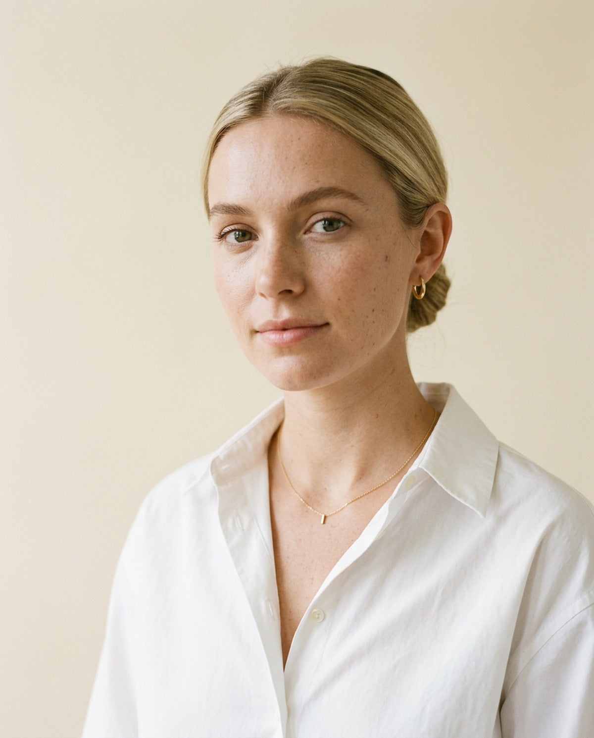 Woman in a crisp white shirt wearing a fine gold bar necklace and small gold huggie earrings