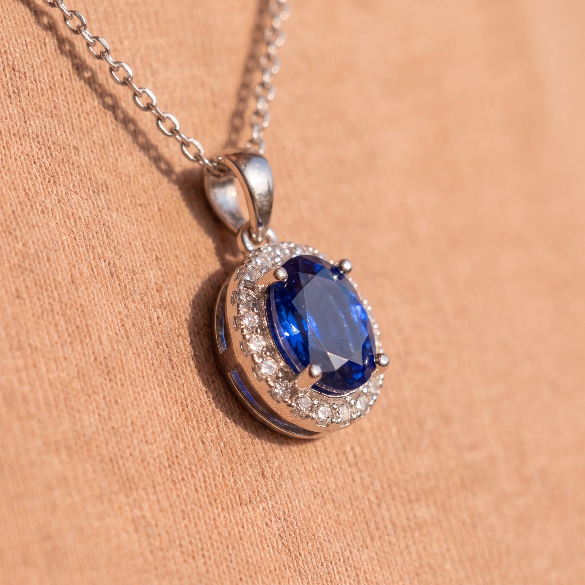 A silver pendant necklace featuring an oval-cut deep blue gemstone surrounded by a halo of small clear stones, resting on a textured peach fabric background.