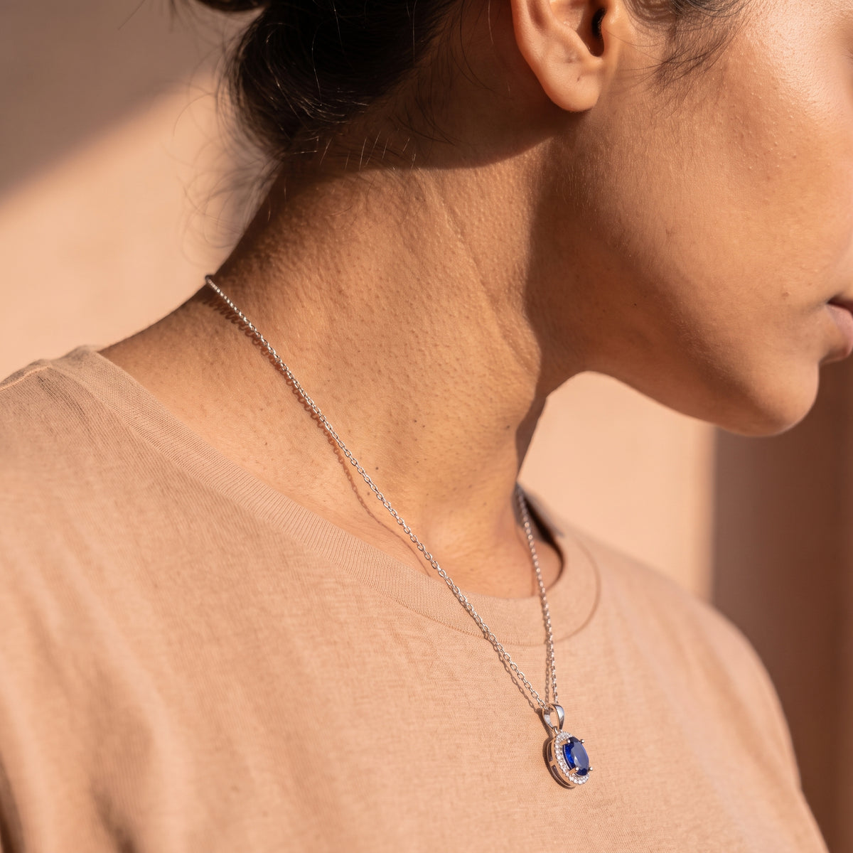 A close-up side profile of a woman wearing a silver chain necklace with a blue oval pendant, paired with a beige crewneck top.