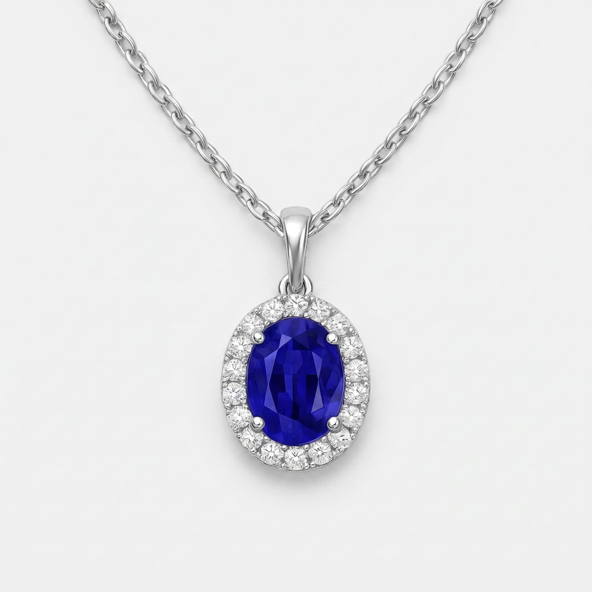 A silver-tone oval pendant necklace featuring a deep blue sapphire center stone surrounded by a halo of small clear stones, suspended from a matching silver chain on a light gray background.
