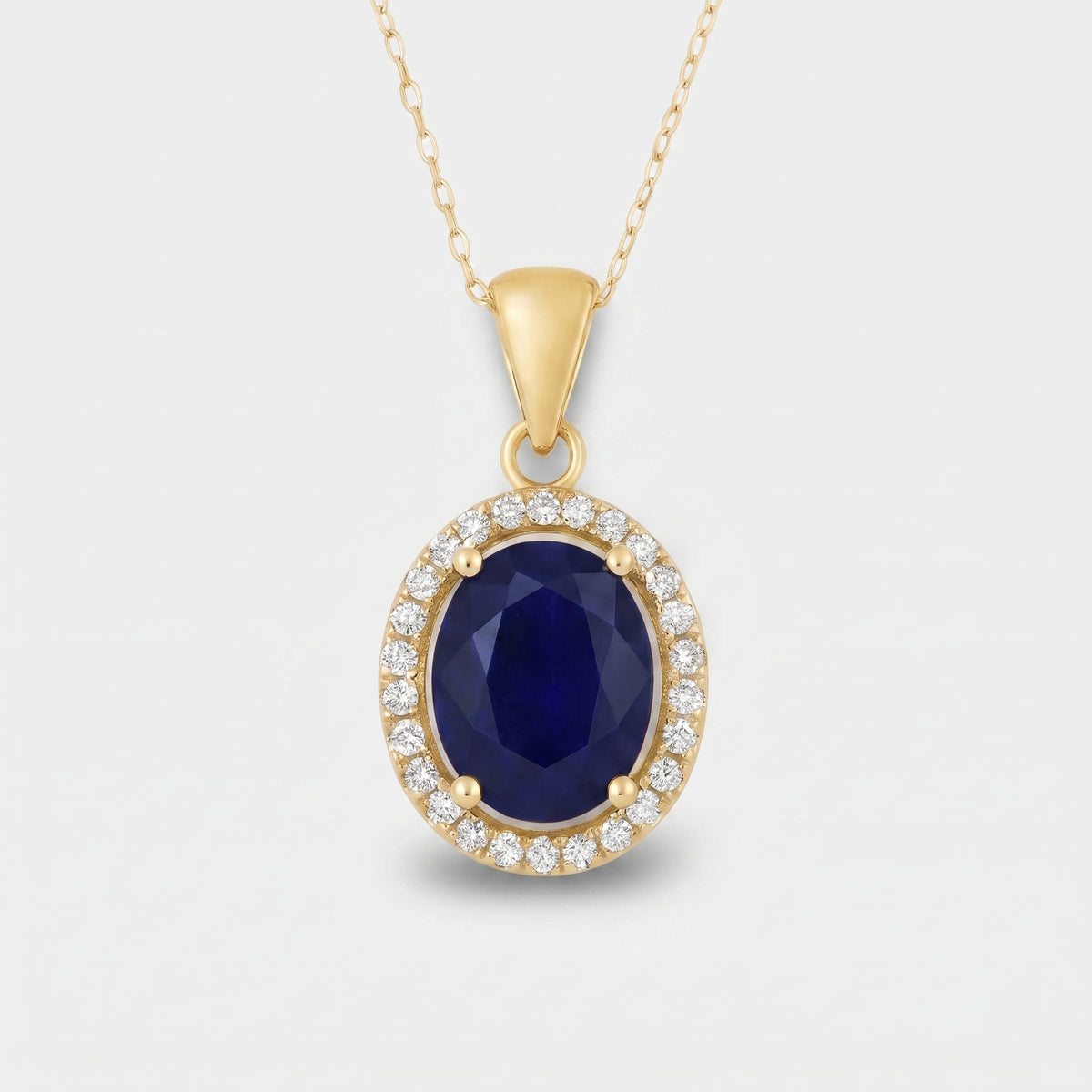 A yellow gold oval pendant necklace featuring a deep blue faceted center stone surrounded by a halo of small clear stones, shown on a light gray background.