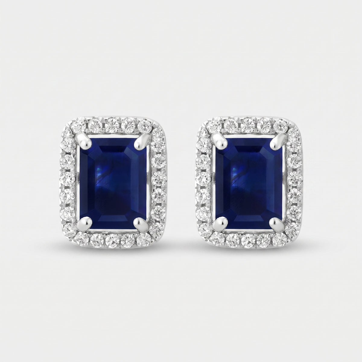 A pair of rectangular stud earrings featuring deep blue sapphires surrounded by a halo of small clear stones in a silver-toned setting, shown on a light gray background.
