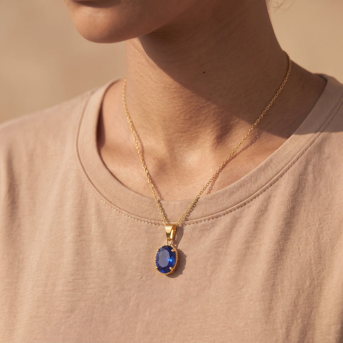 A gold chain necklace with an oval blue gemstone pendant, worn by a person in a beige t-shirt under warm sunlight.