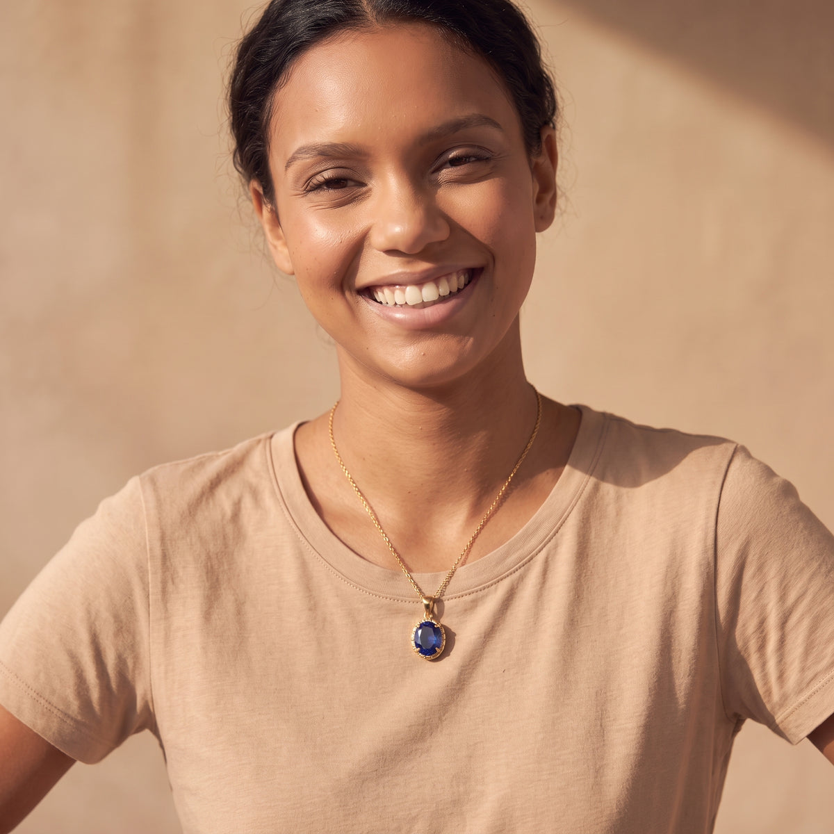 A smiling woman wearing a gold chain necklace with a blue oval pendant, paired with a beige t-shirt against a warm beige background.