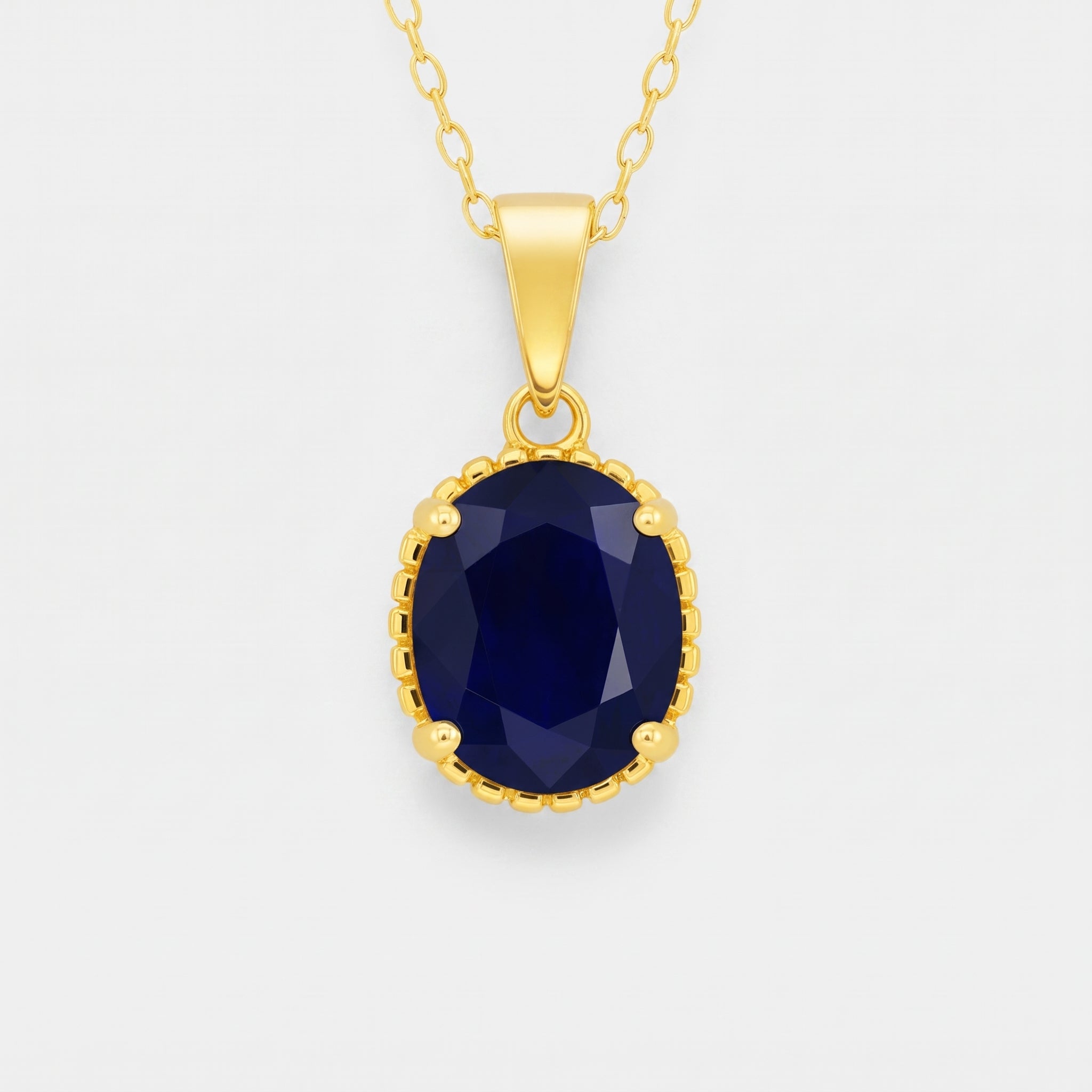 A gold pendant necklace featuring an oval-cut dark blue sapphire gemstone in a beaded gold bezel setting, suspended from a delicate gold chain against a light gray background.