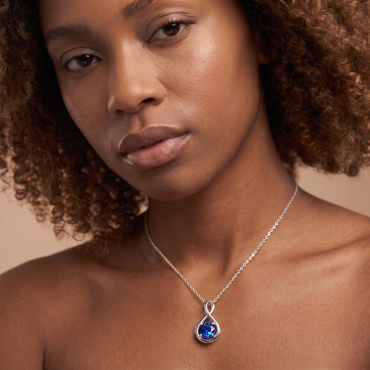 A close-up of a woman wearing a silver chain necklace with a teardrop-shaped blue gemstone pendant, set against a warm beige background.