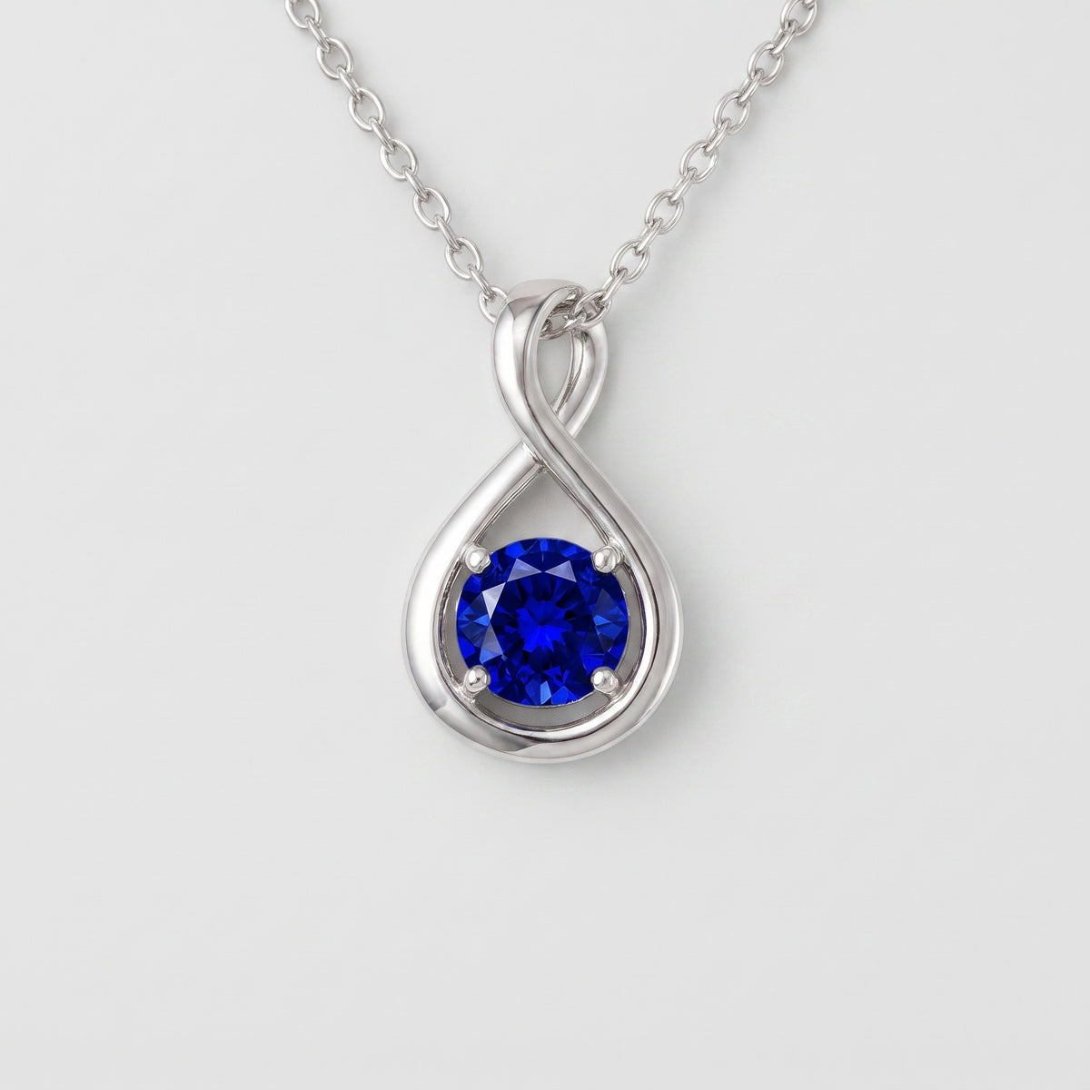 A silver pendant necklace featuring a round-cut deep blue gemstone in a teardrop-shaped setting with an infinity symbol design, suspended from a delicate silver chain against a light gray background.