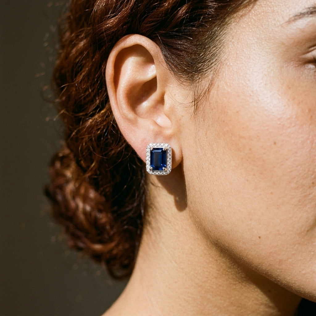 A close-up side profile of a woman wearing a single rectangular blue sapphire stud earring with a halo of small clear stones, set against a dark neutral background.