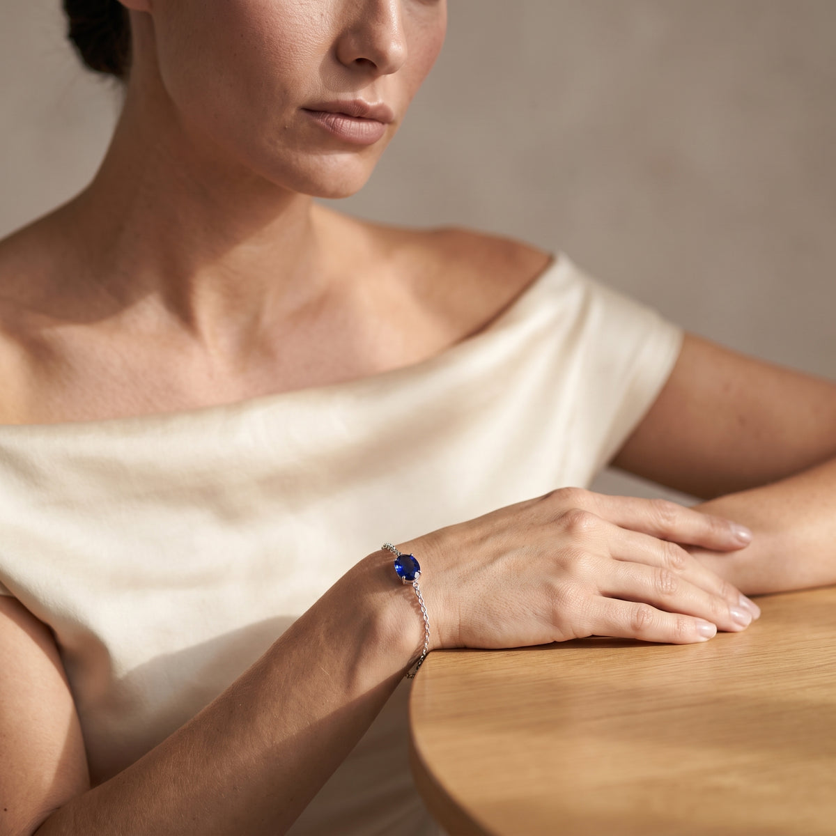 A close-up lifestyle shot of a woman wearing a delicate silver chain bracelet with a deep blue gemstone, resting her hand on a wooden table.
