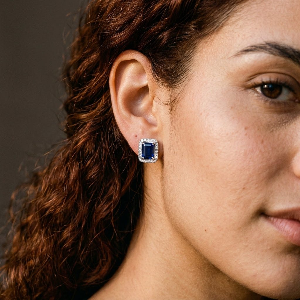 A close-up of a woman's ear wearing a rectangular stud earring with a deep blue center stone surrounded by small clear stones in a silver-toned setting.