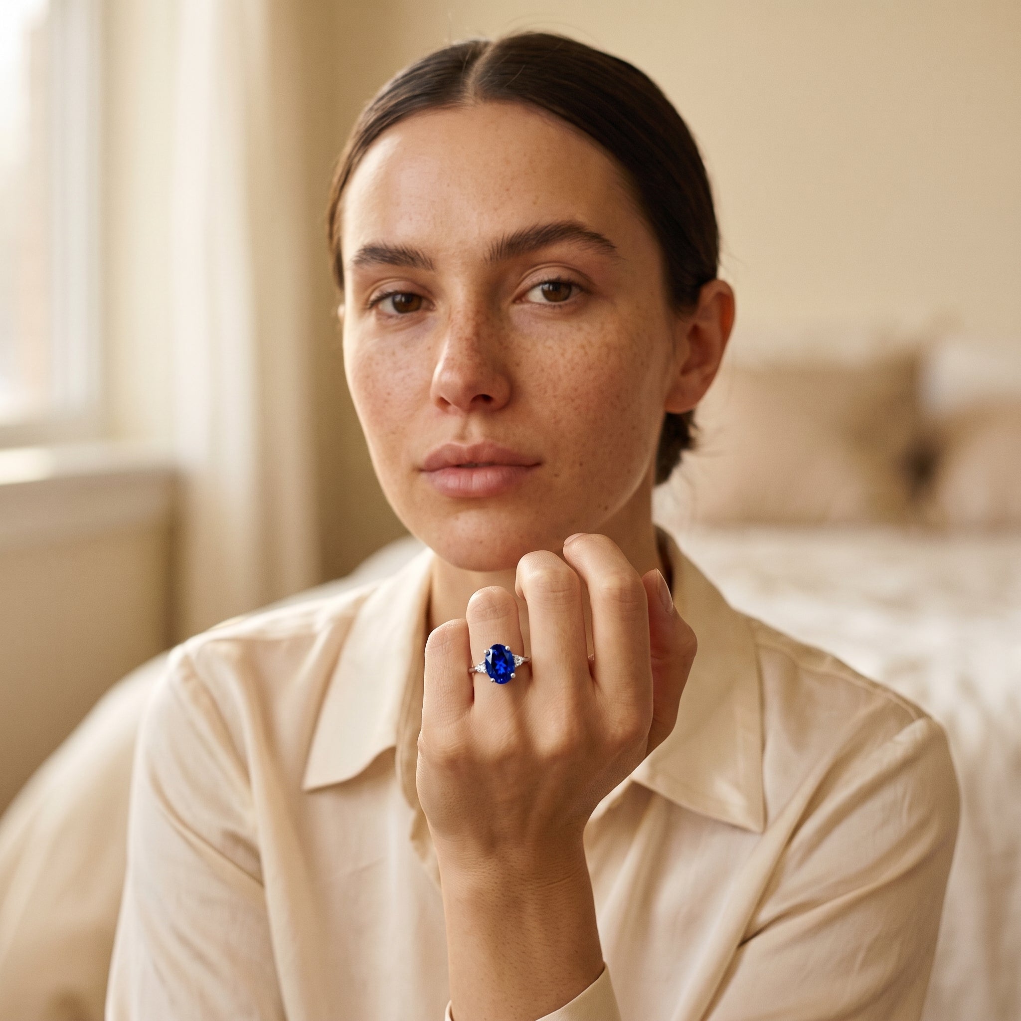 A woman wearing a cream-colored collared shirt and a cream jacket, holding up a ring with a large oval blue gemstone on her finger, set against a softly blurred bedroom background.
