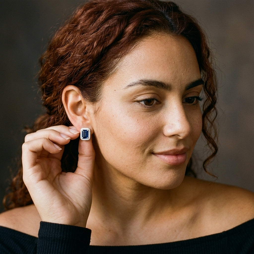 A close-up portrait of a woman with reddish-brown hair wearing a small rectangular stud earring with a dark blue stone and a silver-toned setting, holding the earring with her hand.