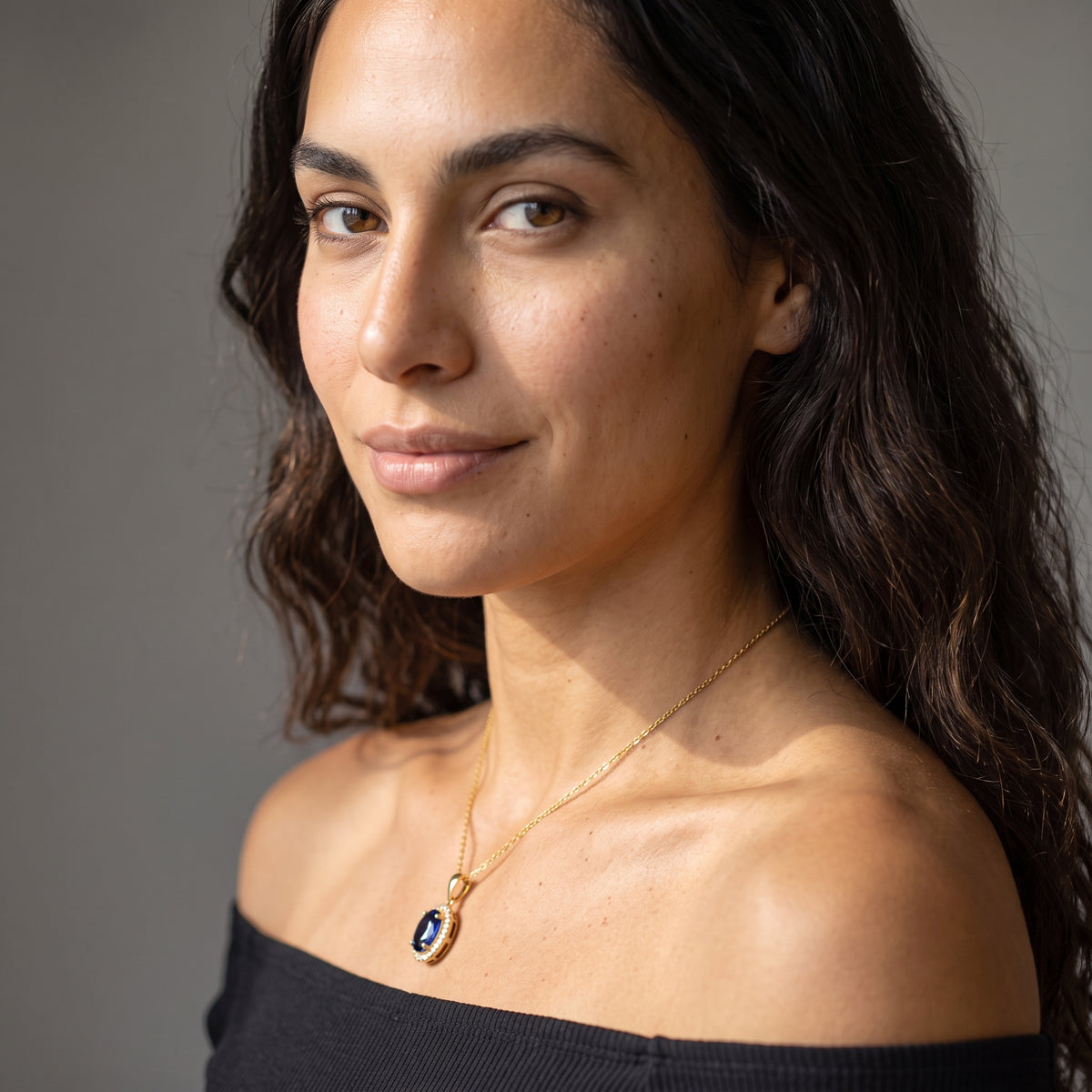 A woman with long dark hair wearing a delicate gold chain necklace with a dark oval pendant, photographed in warm natural light against a neutral gray background.