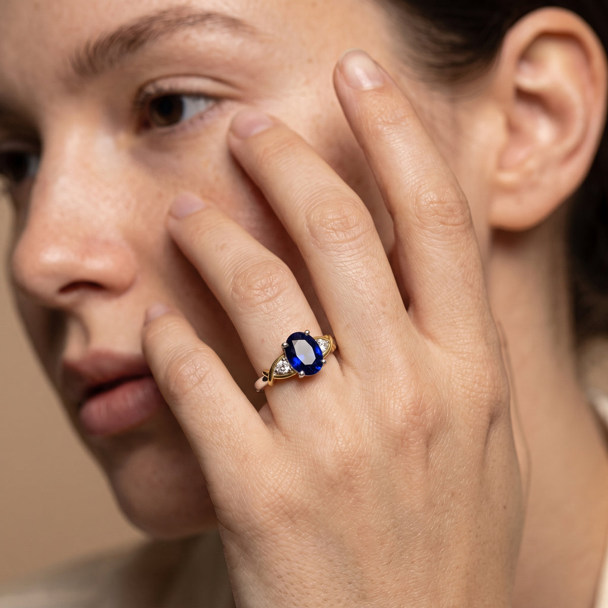 A close-up lifestyle shot of a woman wearing a gold ring with a deep blue oval gemstone and small clear accent stones on her finger.