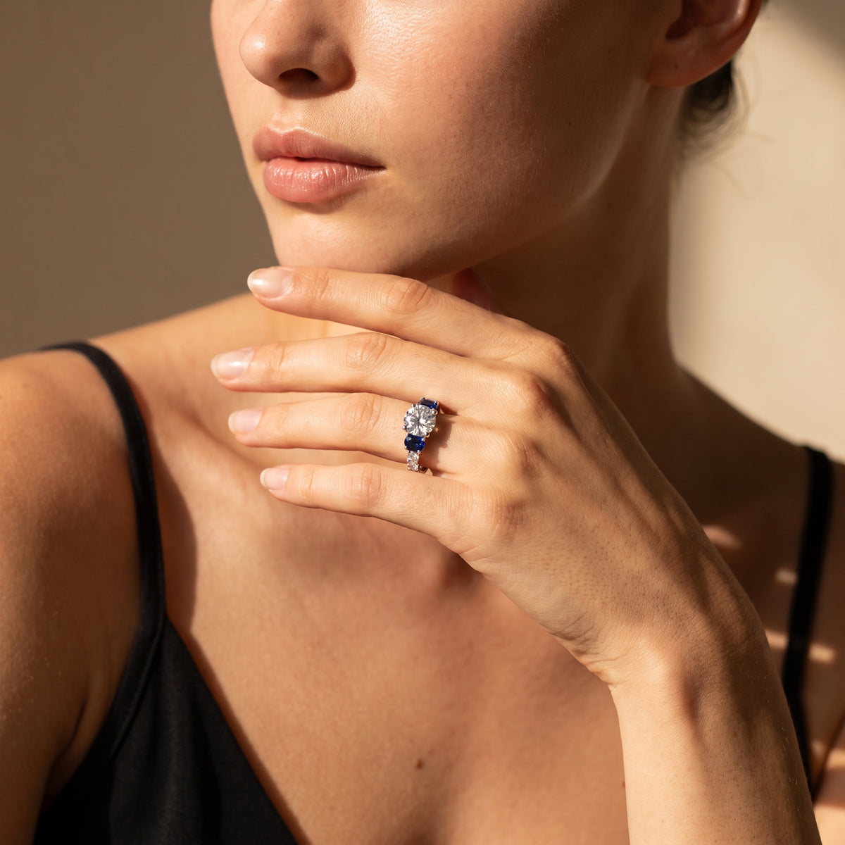 A close-up lifestyle shot of a model wearing a ring with a blue gemstone cluster on a silver-toned band, set against a warm beige background.