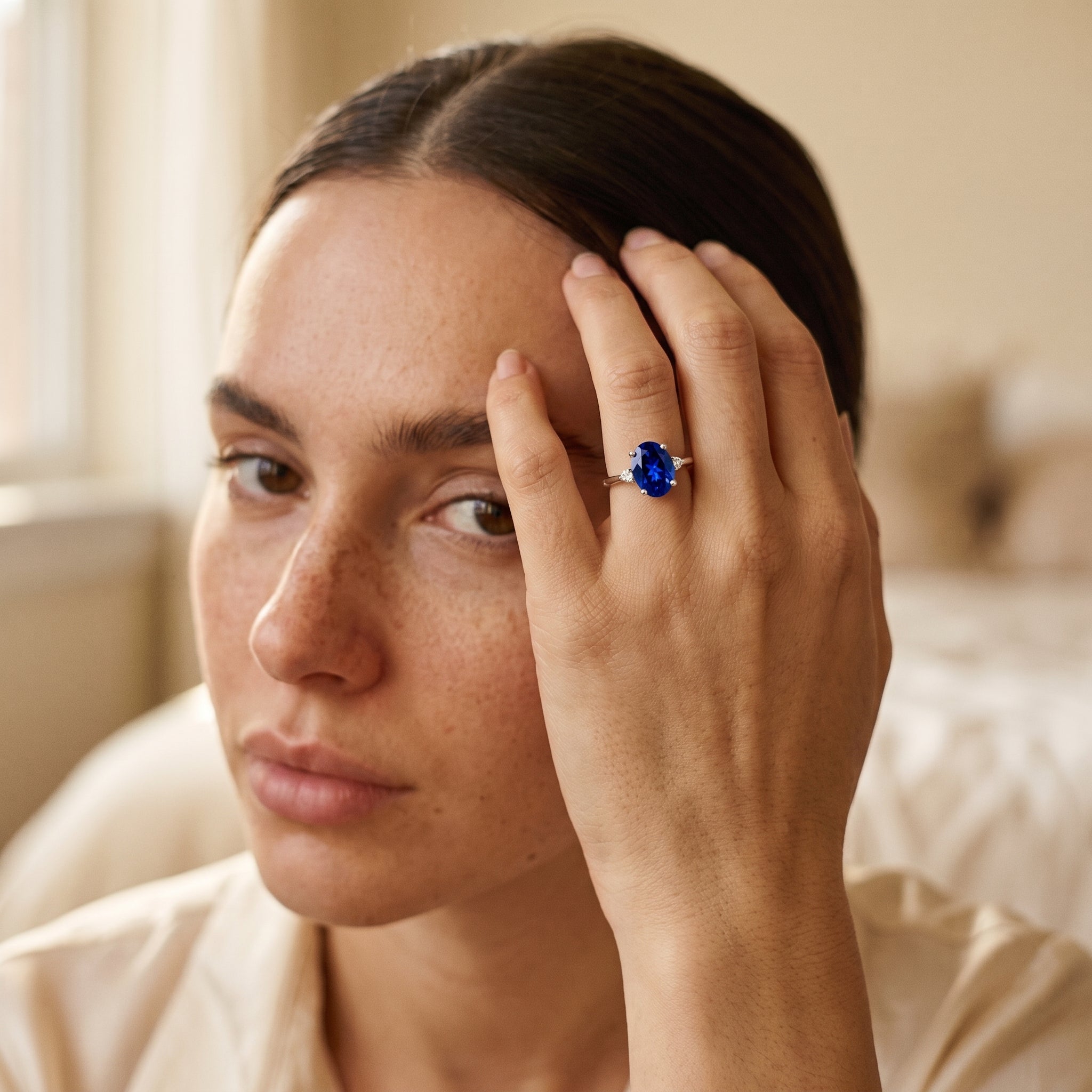 A close-up lifestyle shot of a woman wearing a silver ring with a large oval-cut deep blue gemstone, resting her hand on her head.