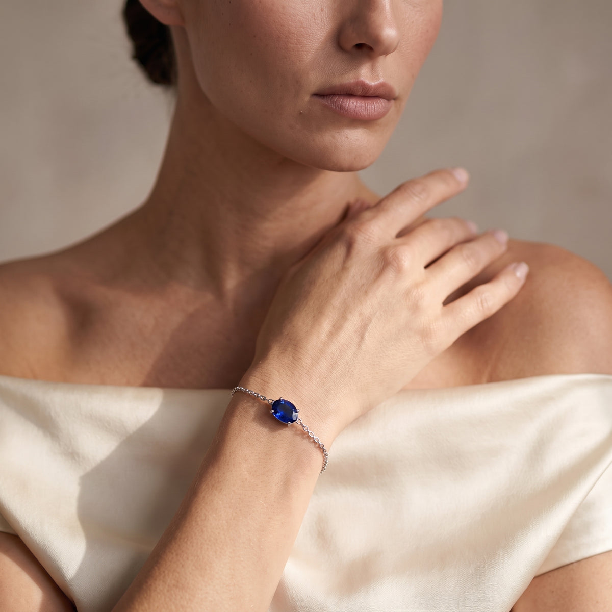 A close-up lifestyle shot of a model wearing a delicate silver chain bracelet with a round, deep blue gemstone centerpiece.