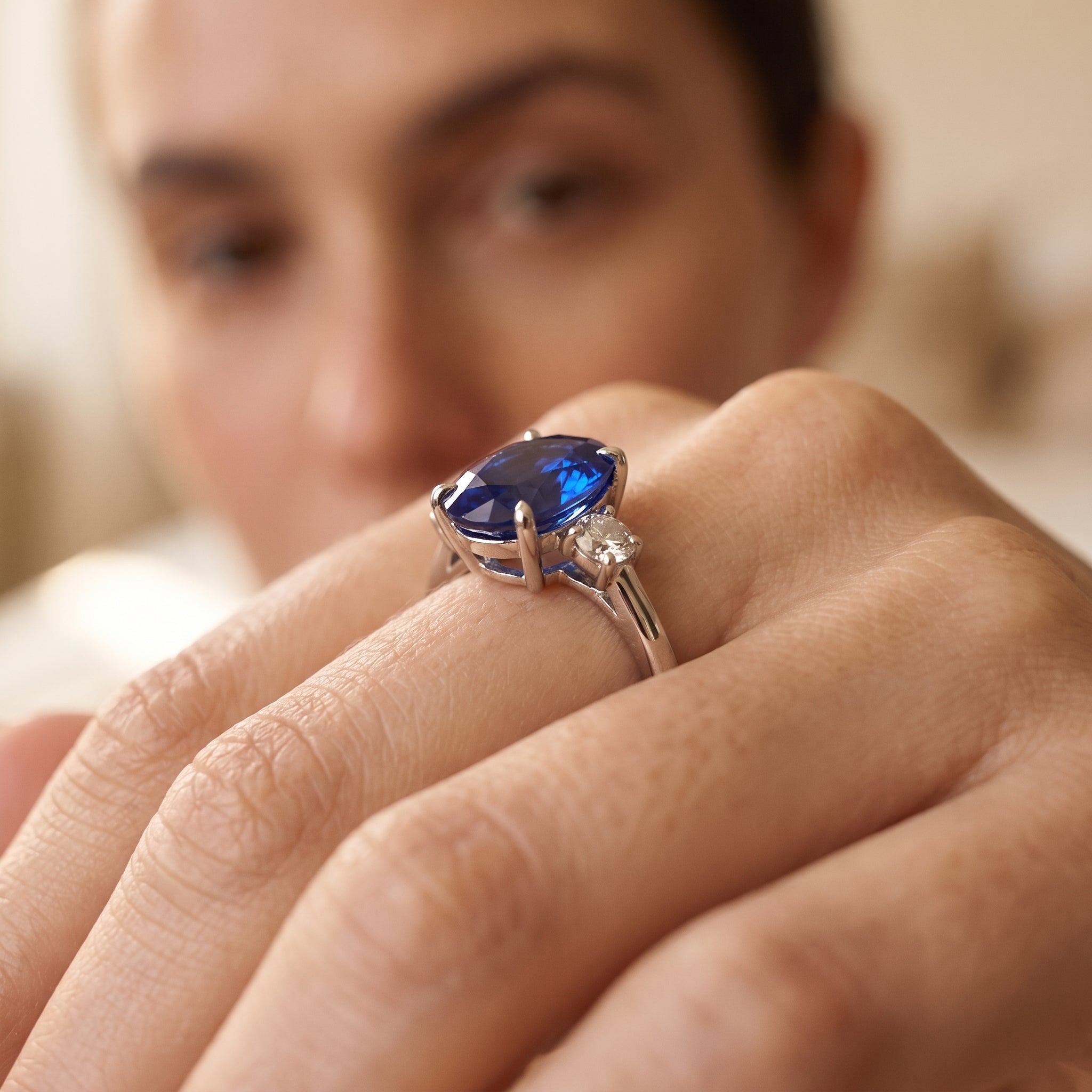 A close-up of a hand wearing a silver ring with a large oval-cut blue gemstone and a smaller clear accent stone, with a softly blurred face in the background.