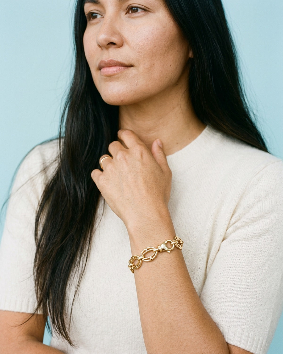 Woman in a cream knit top with her hand at her collarbone, wearing a gold link chain bracelet
