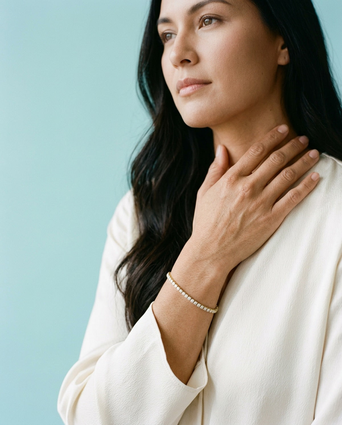 Woman with long dark hair in an ivory blouse, hand at her collarbone, wearing a slim gold diamond tennis bracelet