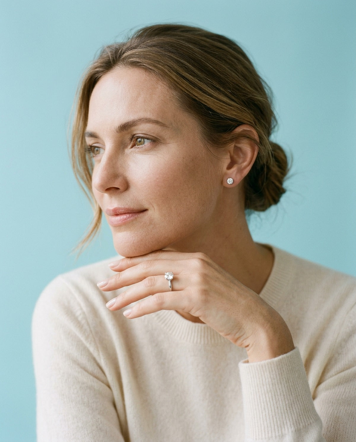 Woman with her hair in a low bun and a cream sweater, chin on her hand, wearing a silver solitaire ring