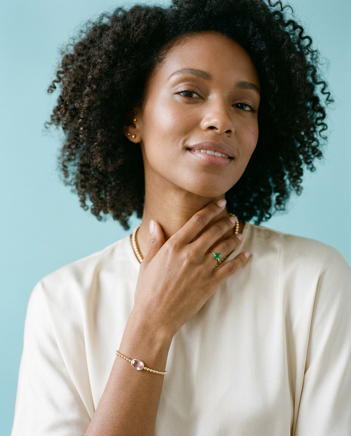 Woman with natural curly hair in a cream top, wearing a gold beaded bracelet with a pink oval stone and a green stone ring