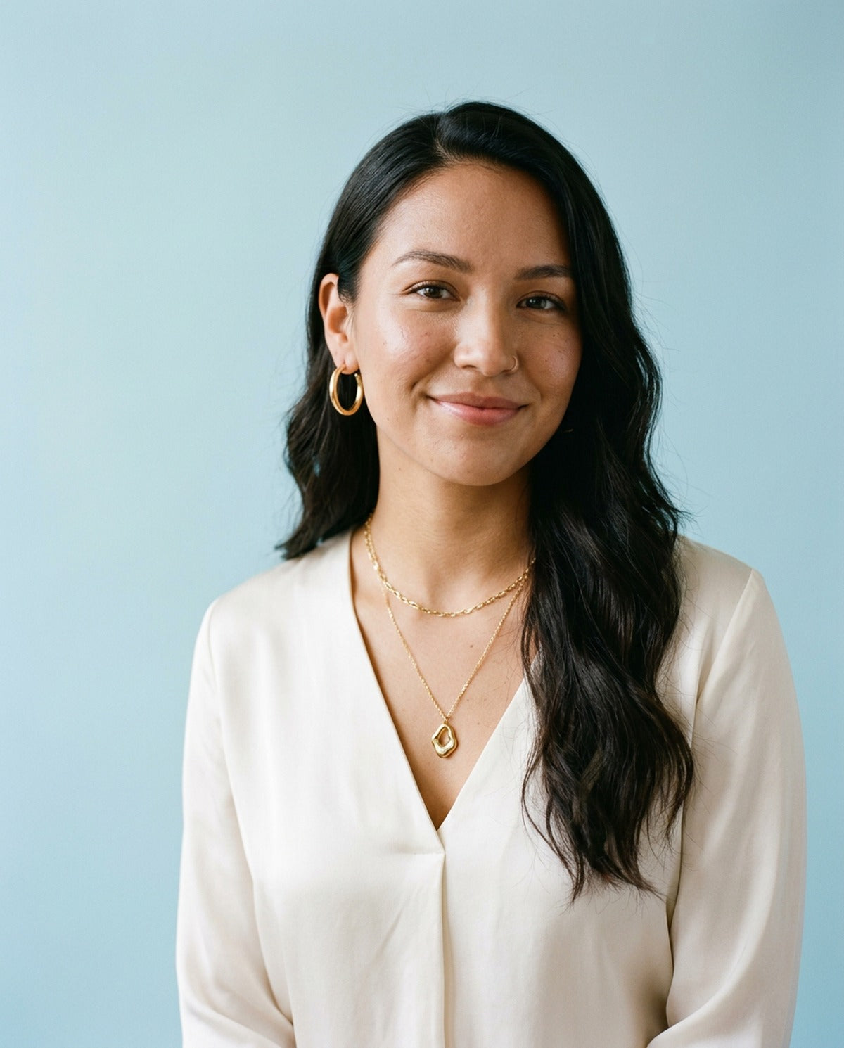 Woman in an ivory silk blouse wearing layered gold chain necklaces and polished gold hoop earrings