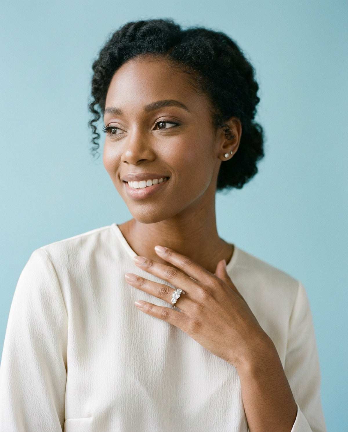 Woman with natural hair in an ivory top, hand at her collarbone, wearing a silver three stone ring