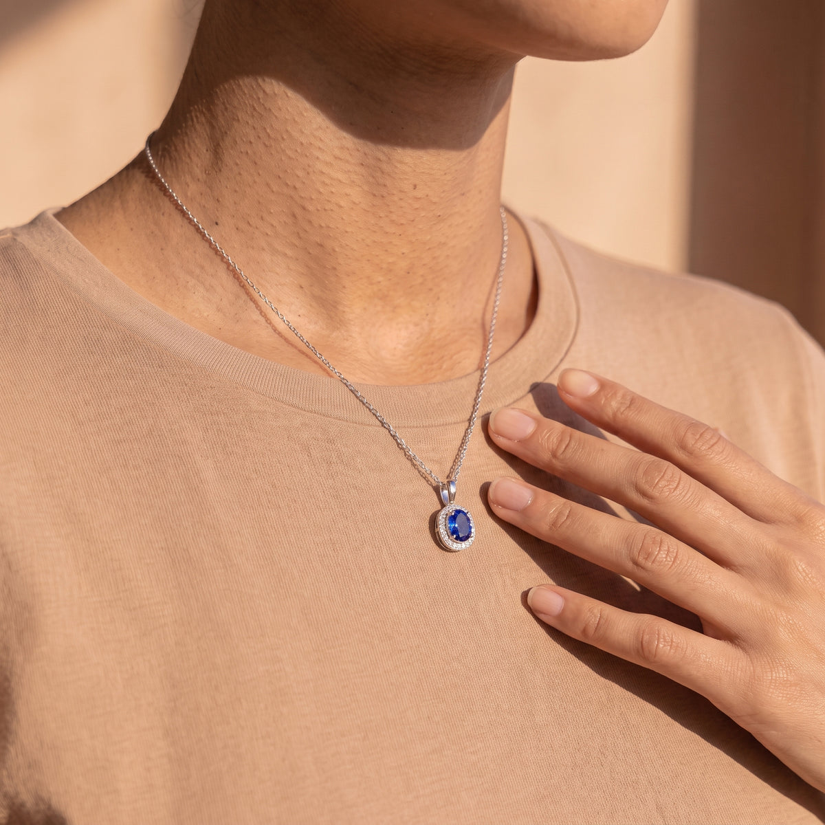 A close-up lifestyle shot of a model wearing a silver chain necklace with a round-cut blue gemstone pendant surrounded by a halo of small clear stones, styled with a beige t-shirt.