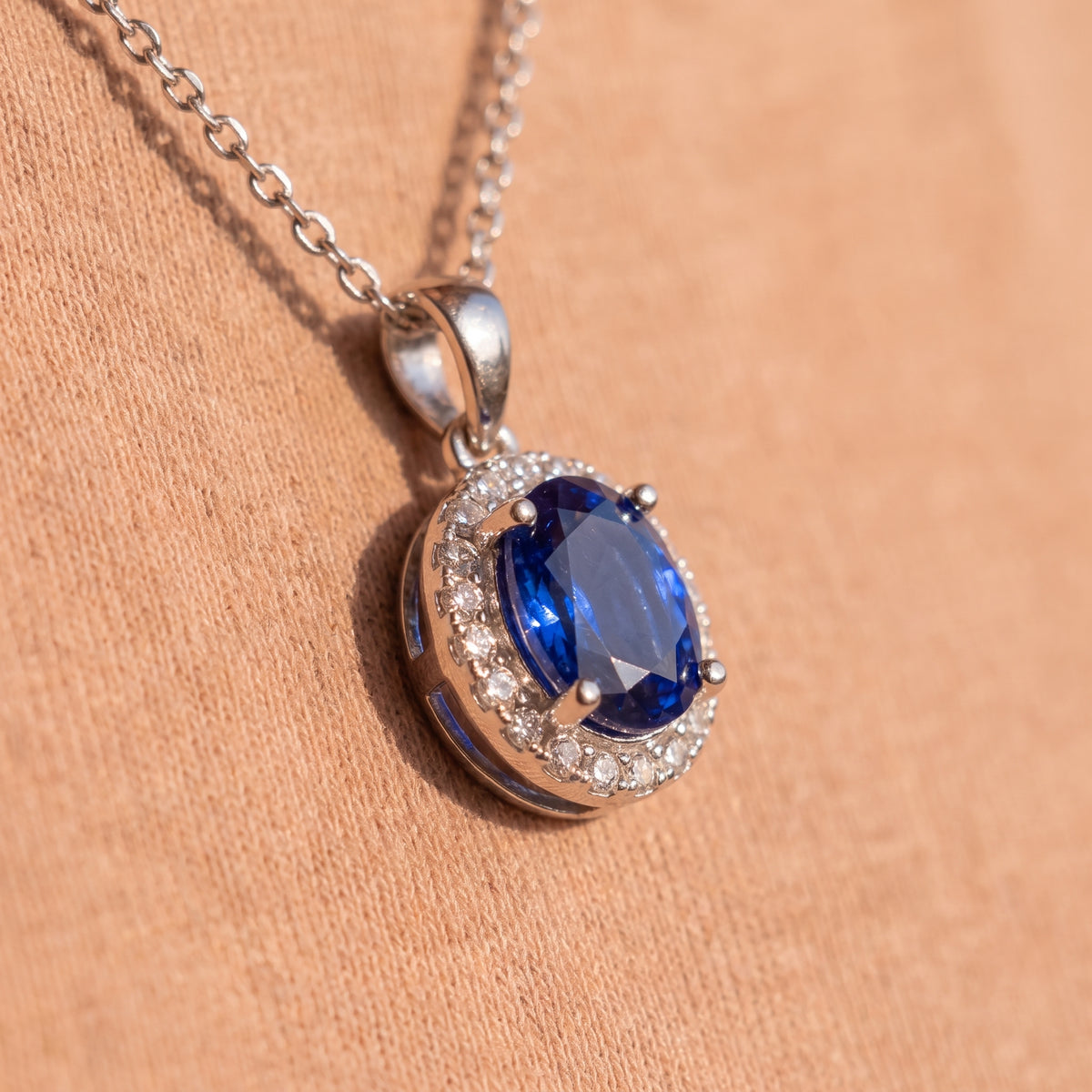 A silver pendant necklace featuring an oval-cut deep blue gemstone surrounded by a halo of small clear stones, resting on a textured peach fabric background.