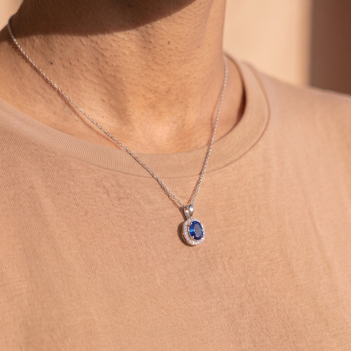 A close-up of a person's neck and upper chest wearing a silver chain necklace with a teardrop-shaped blue gemstone pendant surrounded by a halo of small clear stones, against a beige t-shirt.