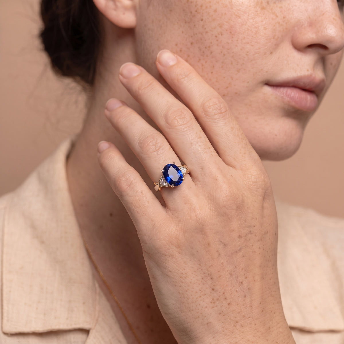 A close-up lifestyle shot of a model wearing a gold ring with a large oval-cut deep blue gemstone. The model is shown from the chest up, wearing a beige linen collared shirt, with her hand resting near her face.