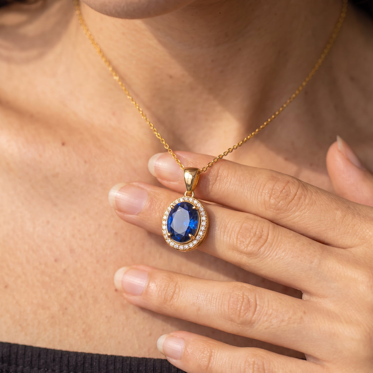 A close-up lifestyle shot of a woman wearing a gold chain necklace with an oval blue sapphire pendant surrounded by a halo of small white stones, resting on her hand.