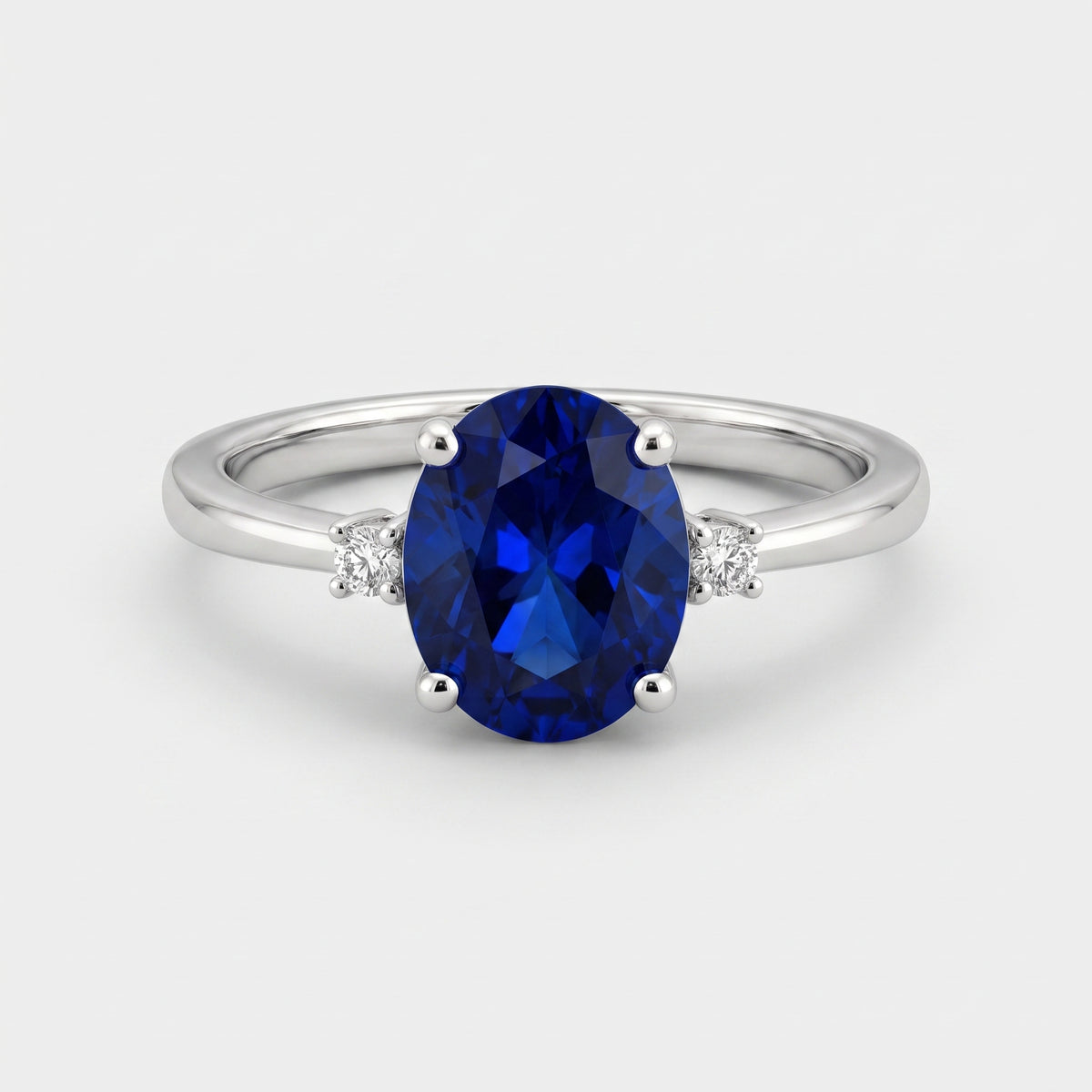 An oval-cut blue sapphire ring set in a polished silver-colored band with small round-cut diamond accents on either side, shown against a clean light gray background.