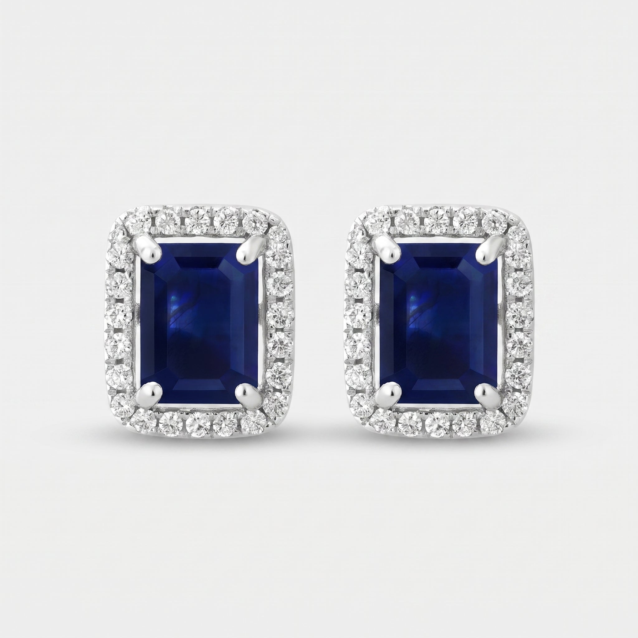 A pair of rectangular stud earrings featuring deep blue sapphires surrounded by a halo of small clear stones in a silver-toned setting, shown on a light gray background.