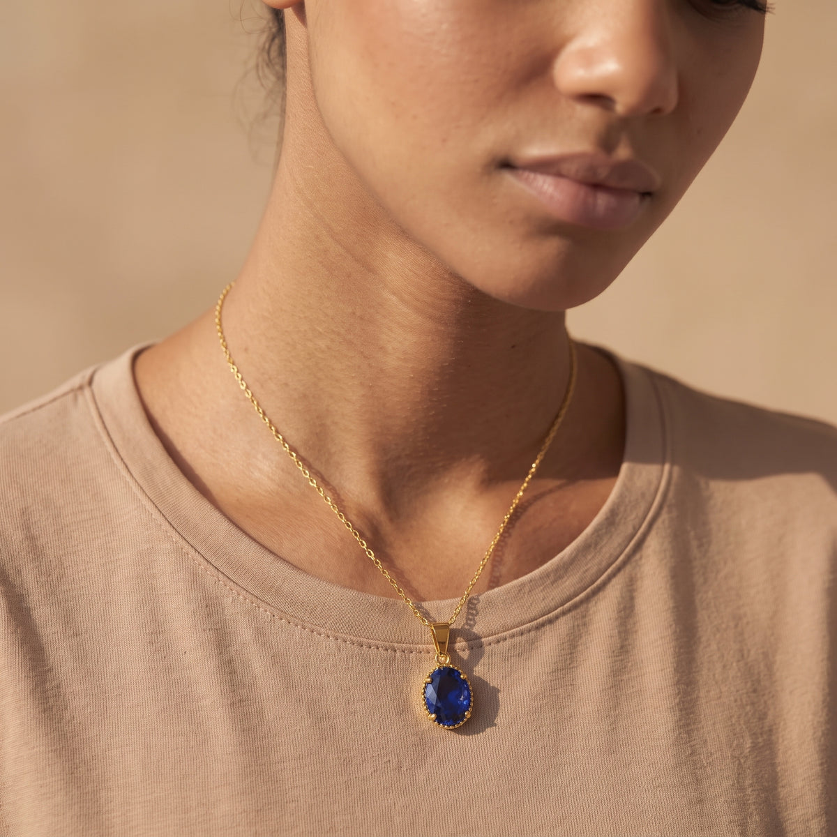 A close-up lifestyle shot of a model wearing a gold chain necklace with a deep blue oval pendant, styled with a beige crew-neck t-shirt against a warm beige background.