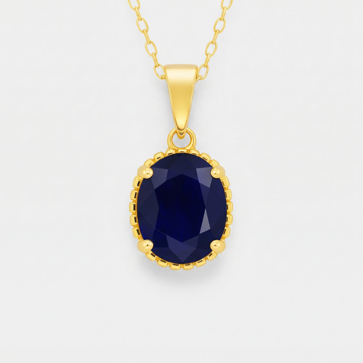 A gold pendant necklace featuring an oval-cut dark blue sapphire gemstone in a beaded gold bezel setting, suspended from a delicate gold chain against a light gray background.