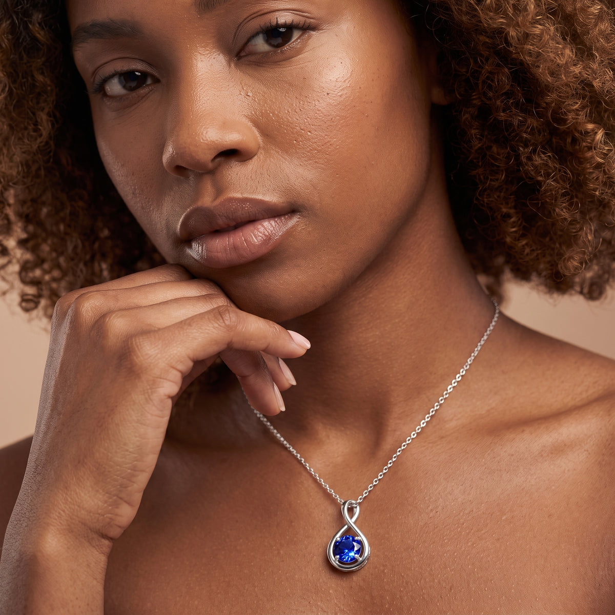 A close-up of a woman wearing a silver teardrop pendant necklace with a deep blue teardrop-cut gemstone, set against a warm beige background.