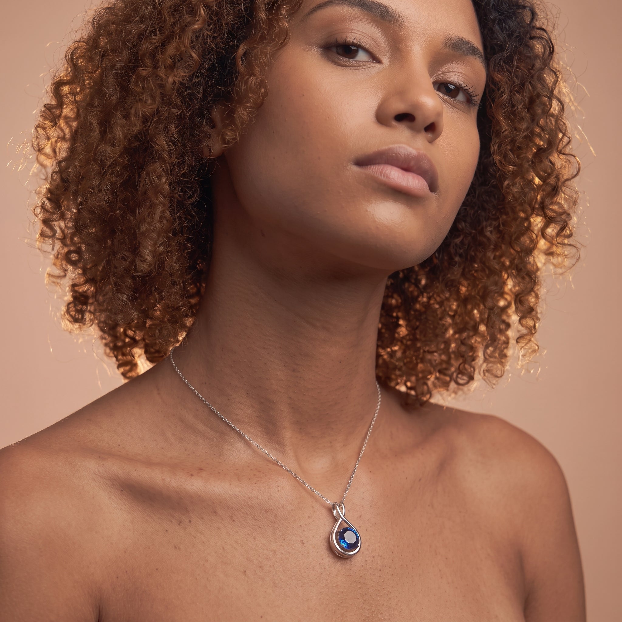A close-up of a woman with curly brown hair wearing a silver teardrop pendant necklace with a blue evil eye design, set against a solid warm beige background.