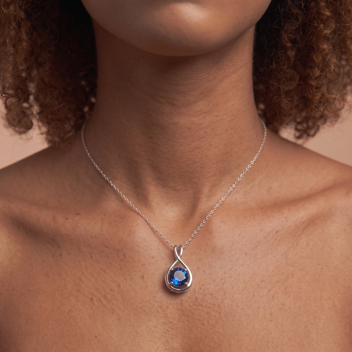 A close-up of a model wearing a delicate silver chain necklace with a teardrop-shaped pendant featuring a deep blue faceted gemstone in a silver bezel setting.