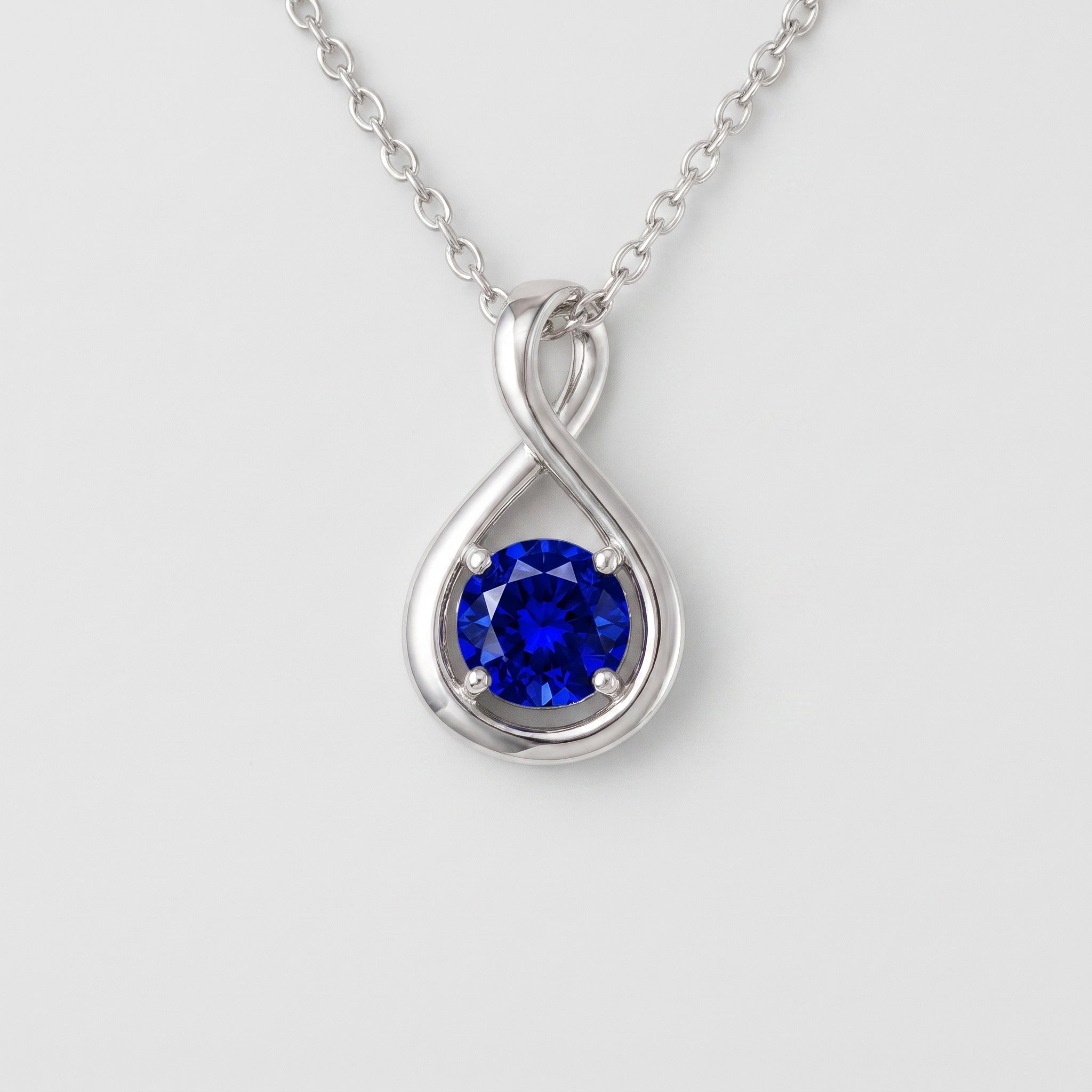 A silver pendant necklace featuring a round-cut deep blue gemstone in a teardrop-shaped setting with an infinity symbol design, suspended from a delicate silver chain against a light gray background.
