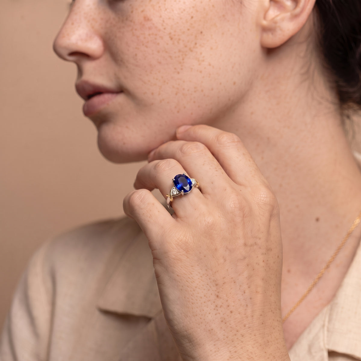 A close-up lifestyle shot of a woman wearing a gold ring with a deep blue oval gemstone and small clear accent stones on her finger, set against a warm beige background.