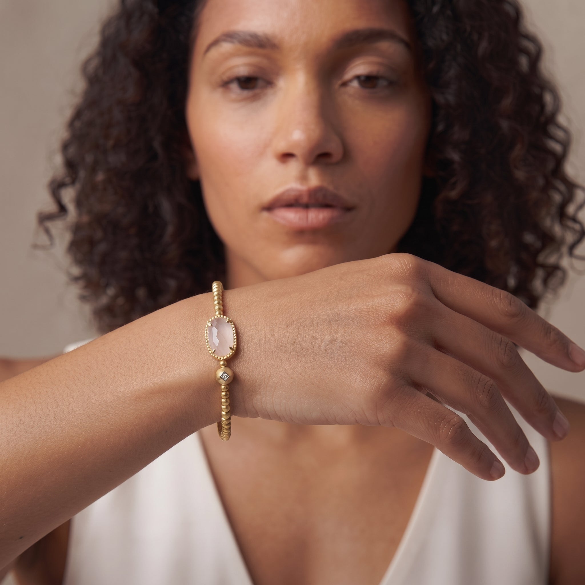A woman wearing a delicate gold chain bracelet with a large oval pale pink stone on her wrist, shown in a close-up portrait against a soft beige background.