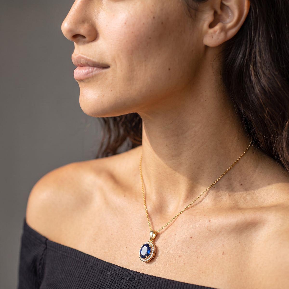 A close-up side profile of a woman wearing a delicate gold chain necklace with a dark blue oval pendant set in a gold bezel.