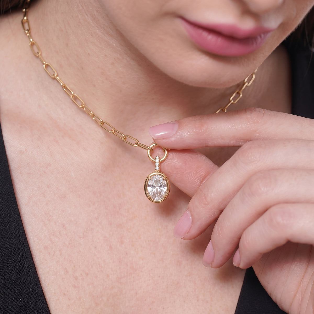 Close-up of a hand holding an oval bezel pendant on a gold paperclip chain at the collarbone