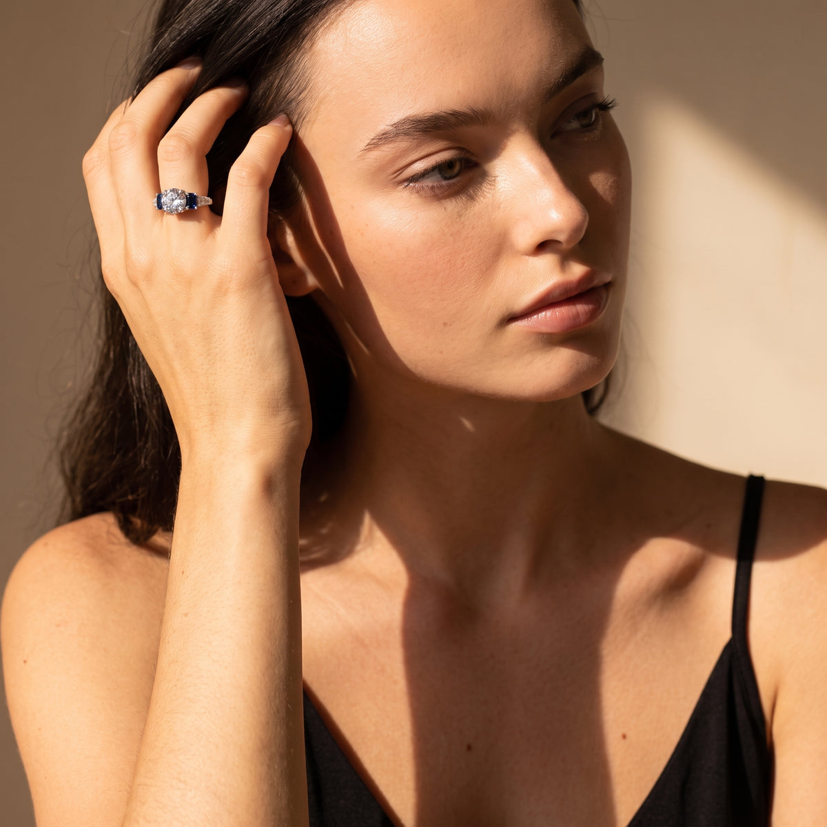 A close-up lifestyle shot of a woman wearing a silver ring with a blue gemstone on her finger, set against a warm beige background with dramatic sunlight and shadows.