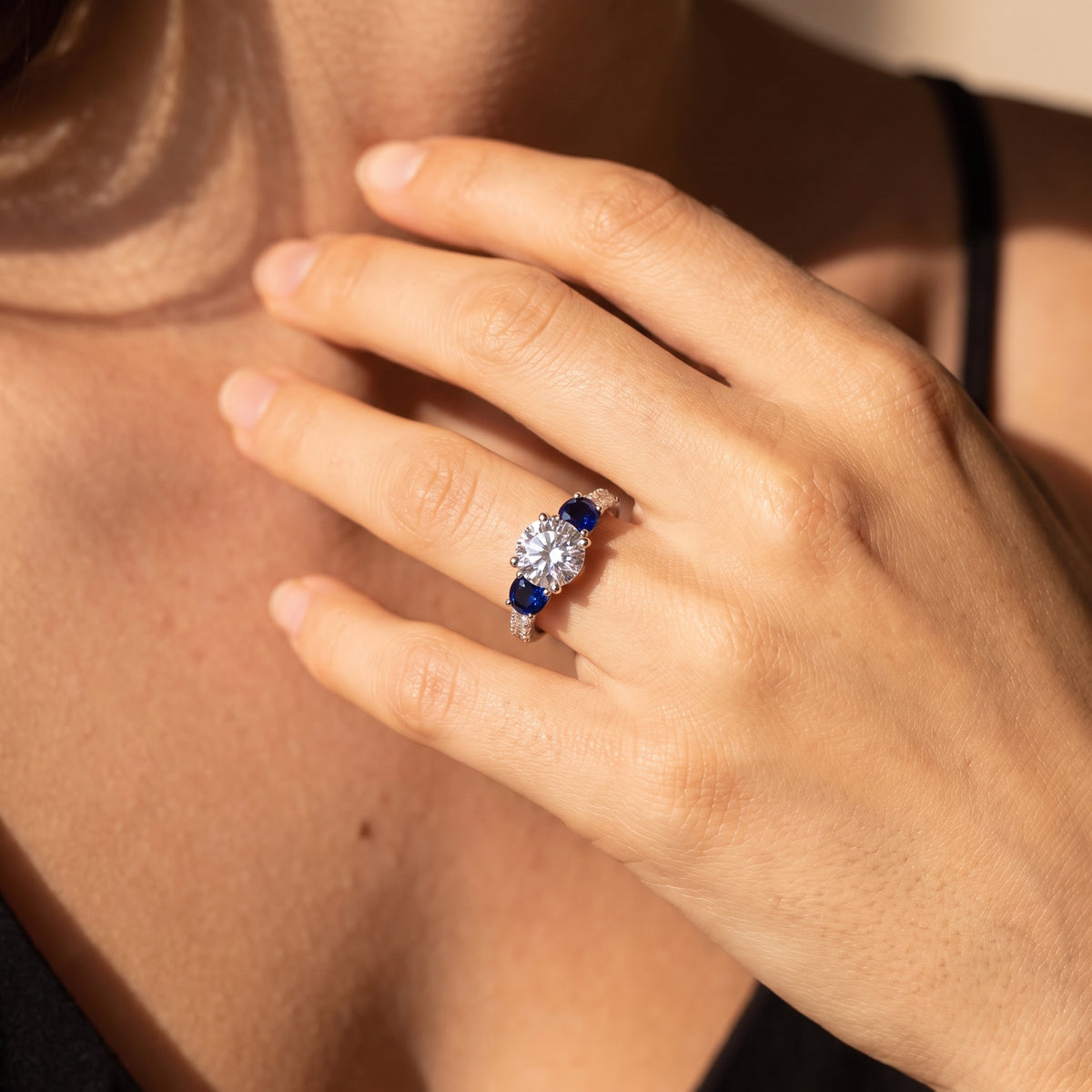 A close-up lifestyle shot of a woman's hand wearing a silver ring with a central round-cut diamond cluster flanked by two oval blue sapphires, set against her neck and shoulder.